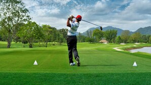 A golf course with a golfer in mid-swing, trees, and a distant mountain. - THE MOMENT (Khlong Luang)