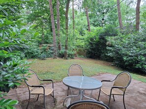 Un patio con una mesa redonda y cuatro sillas de mimbre, rodeado de abundante vegetación y árboles altos.
