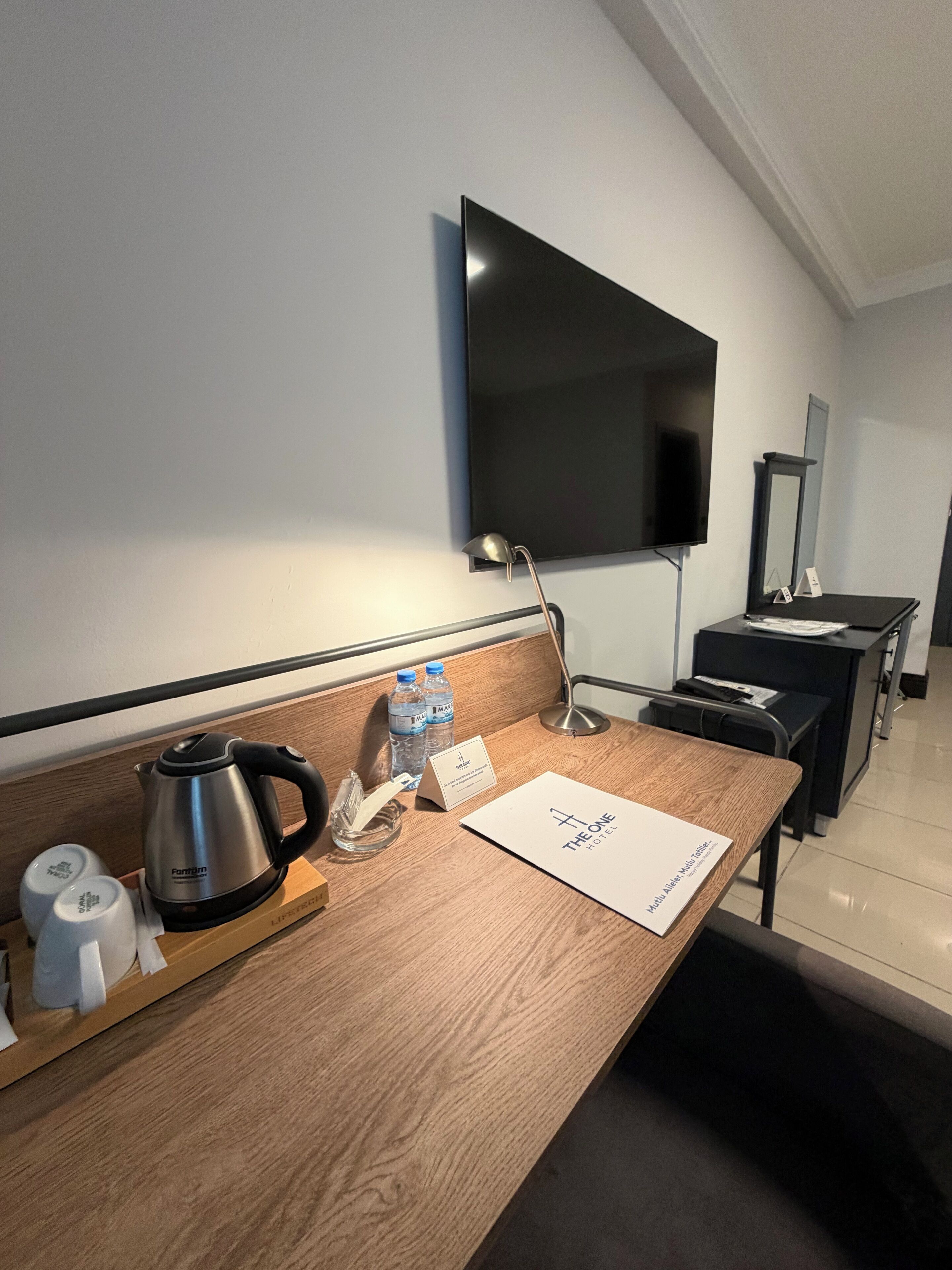 Een hotelkamer met een houten bureau, een waterkoker, flessenwater, een notitieboekje en een flatscreen televisie aan de muur.