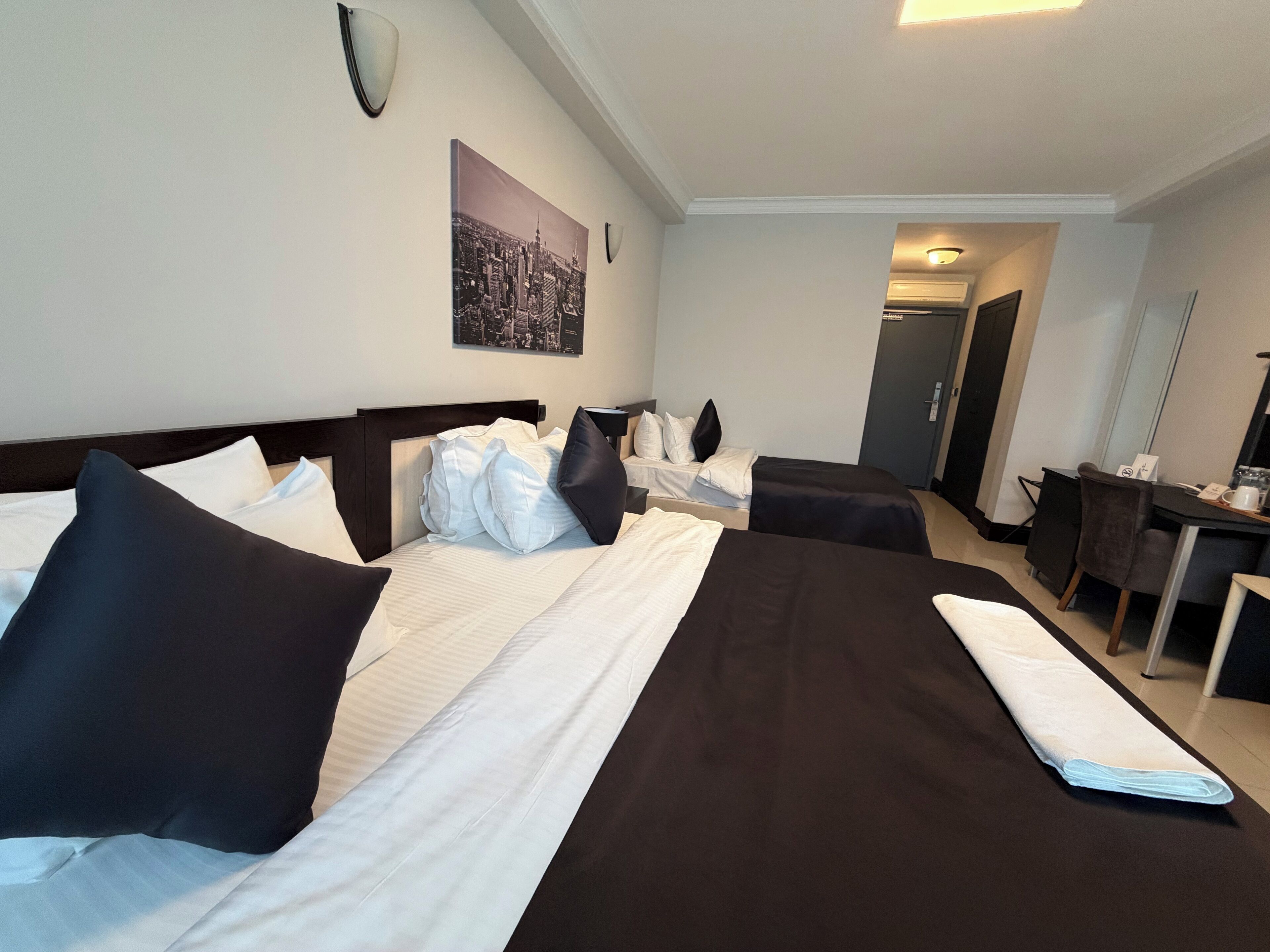 Habitación de hotel con dos camas, un escritorio, una silla, un espejo y una pintura de paisaje urbano en la pared.