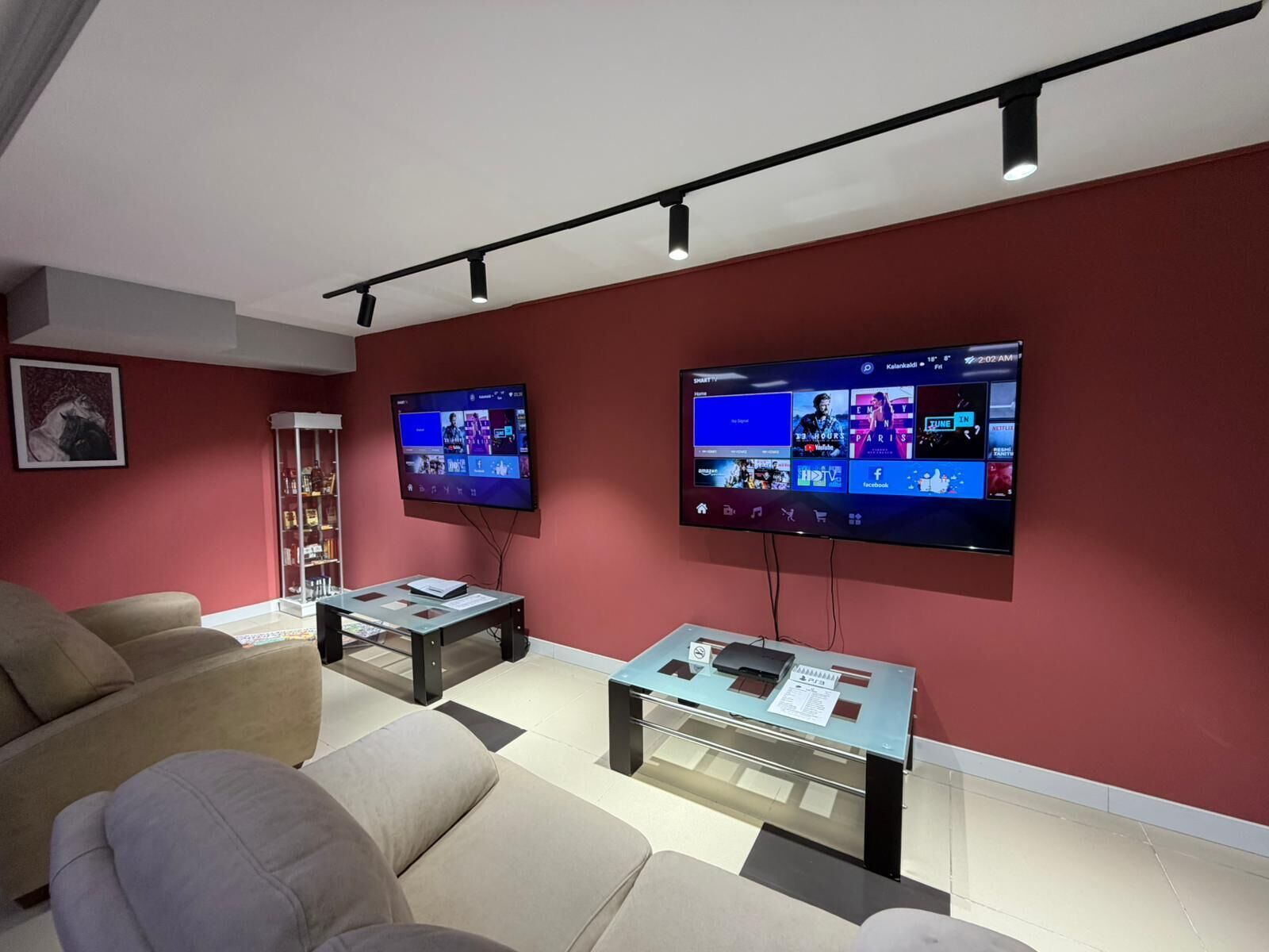 A modern living room with two large flat-screen TVs mounted on the wall, a glass coffee table, a beige sofa, and a red accent wall.