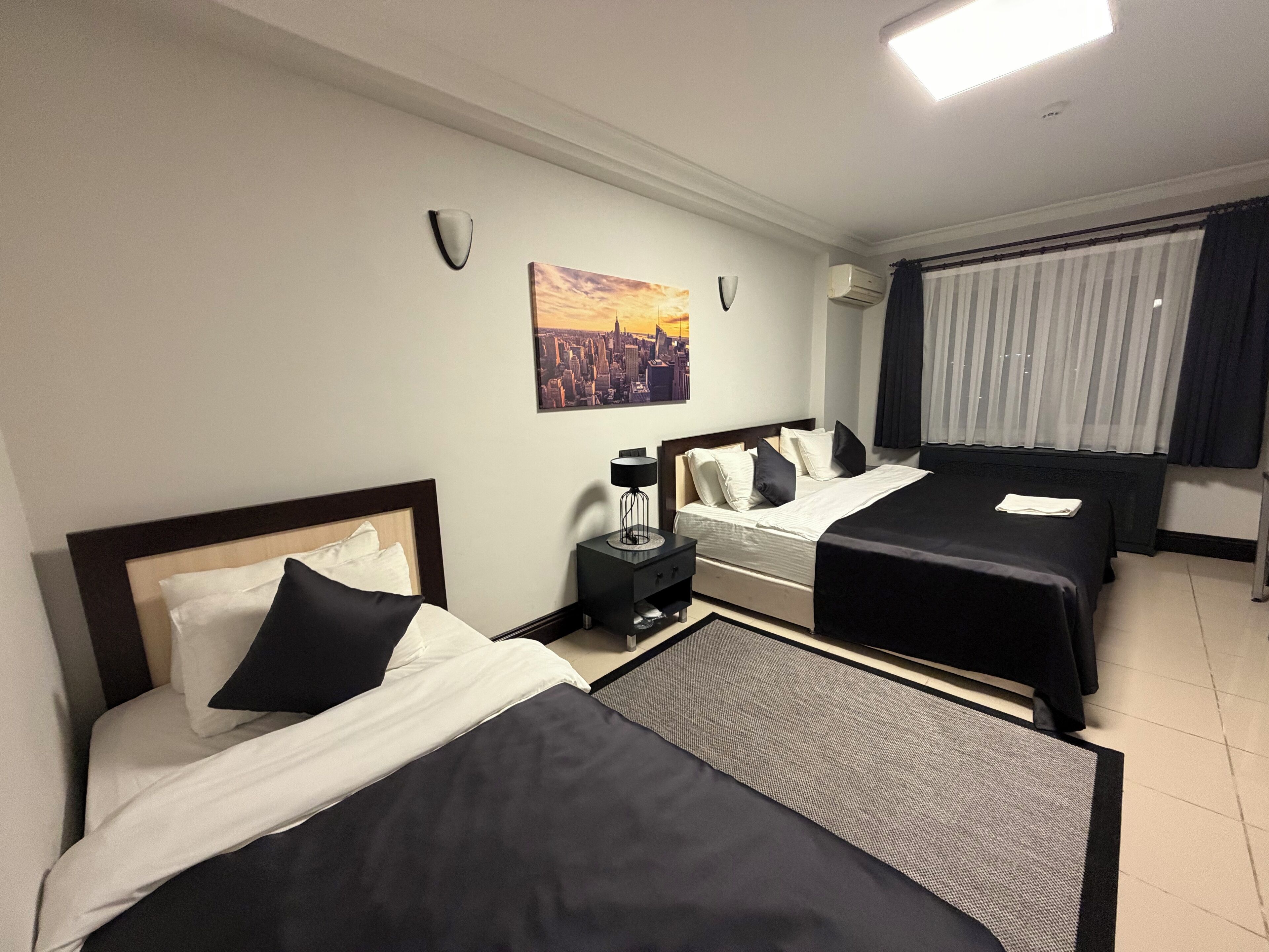 A hotel room with two beds, a nightstand, a lamp, a painting, and a window with curtains.