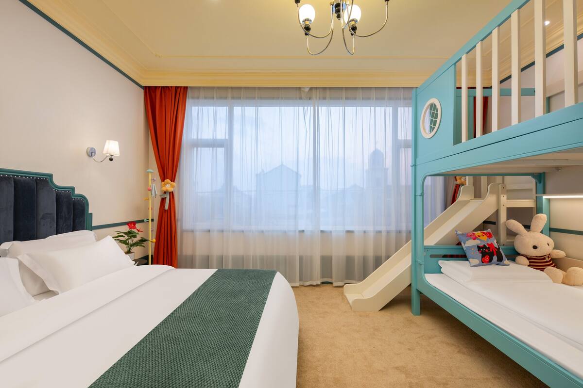 A hotel room with a bed, a bunk bed, a window with curtains, a chandelier, and a nightstand with a lamp.