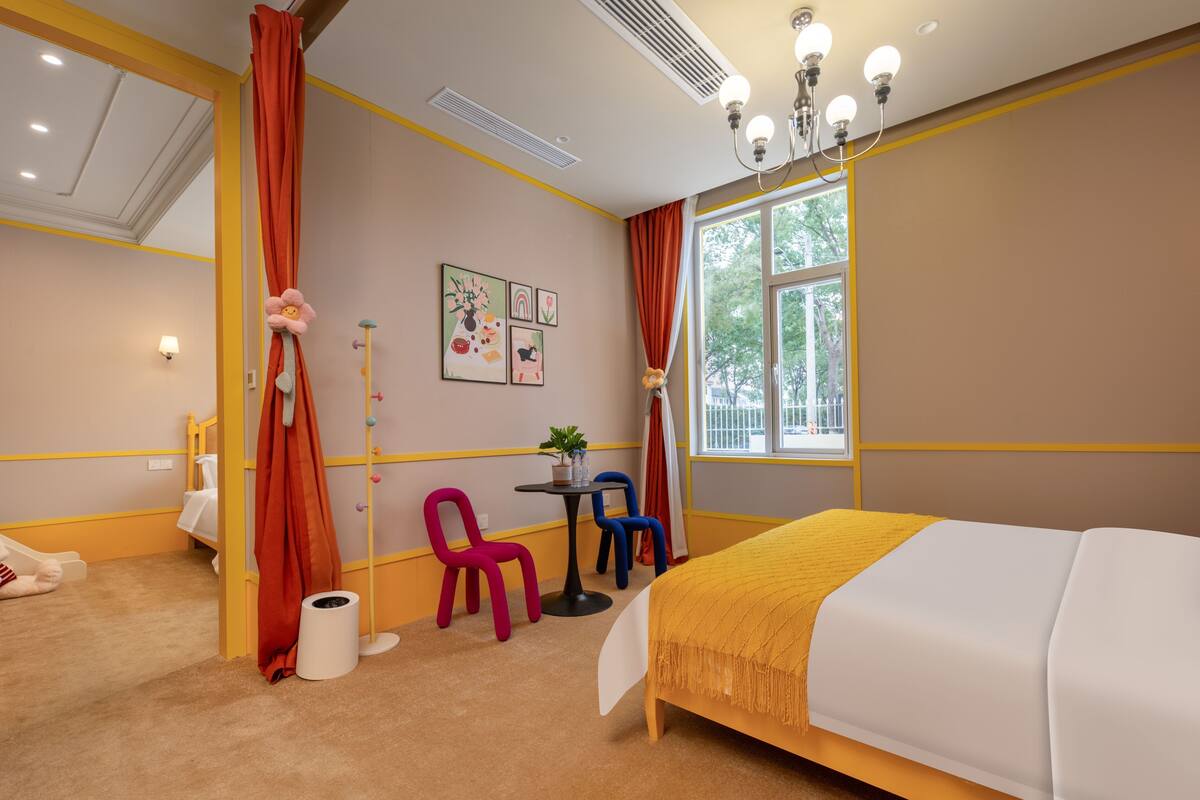 A hotel room with a bed, a small table with chairs, a window with curtains, and a wall with framed pictures.