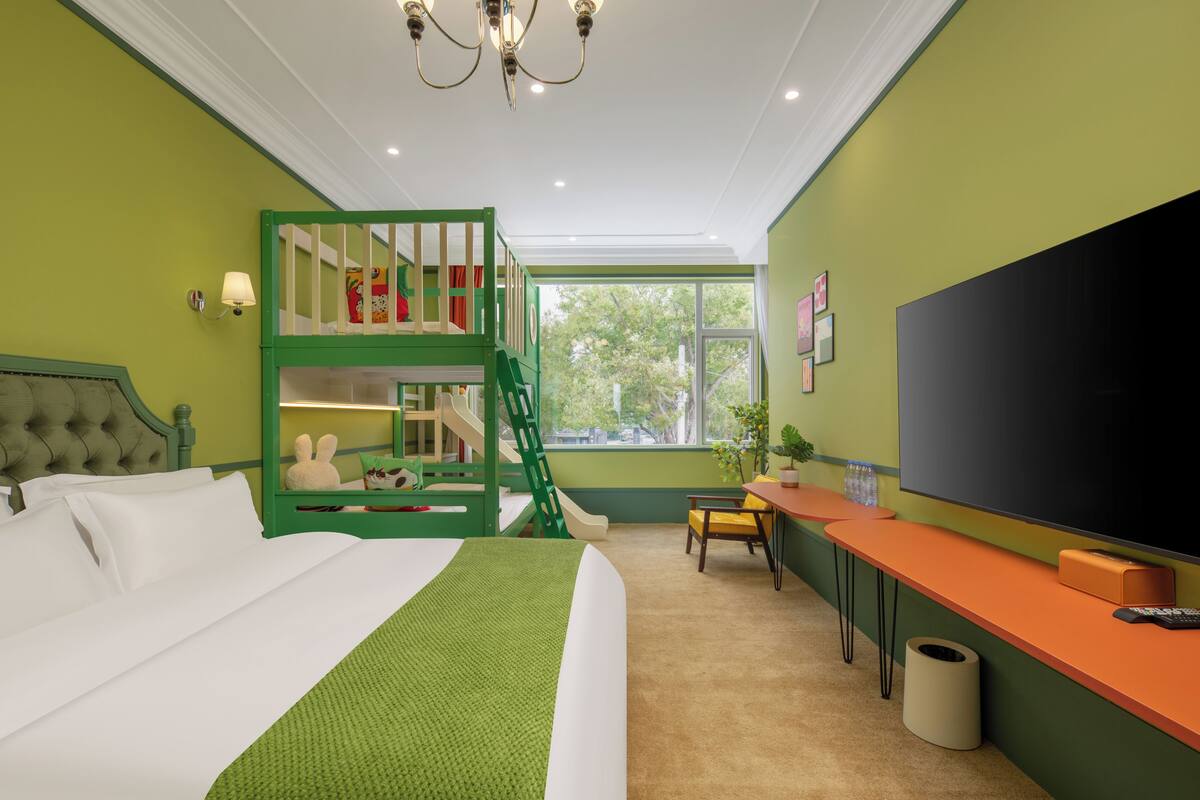 A hotel room with a bed, a green bunk bed, a desk with a TV, and a window with a view of trees.