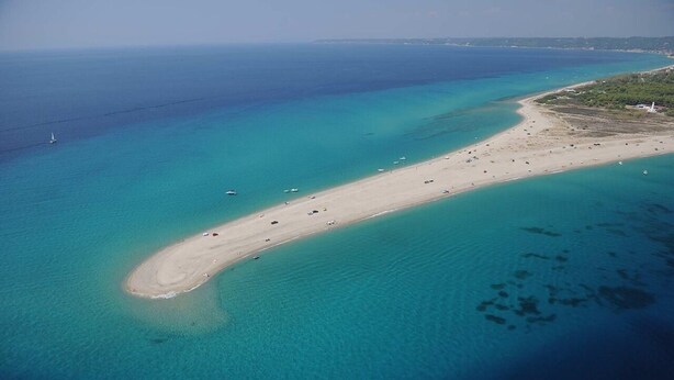 An aerial view of a beach with clear turquoise waters, a sandy shore, and a lighthouse in the distance.
