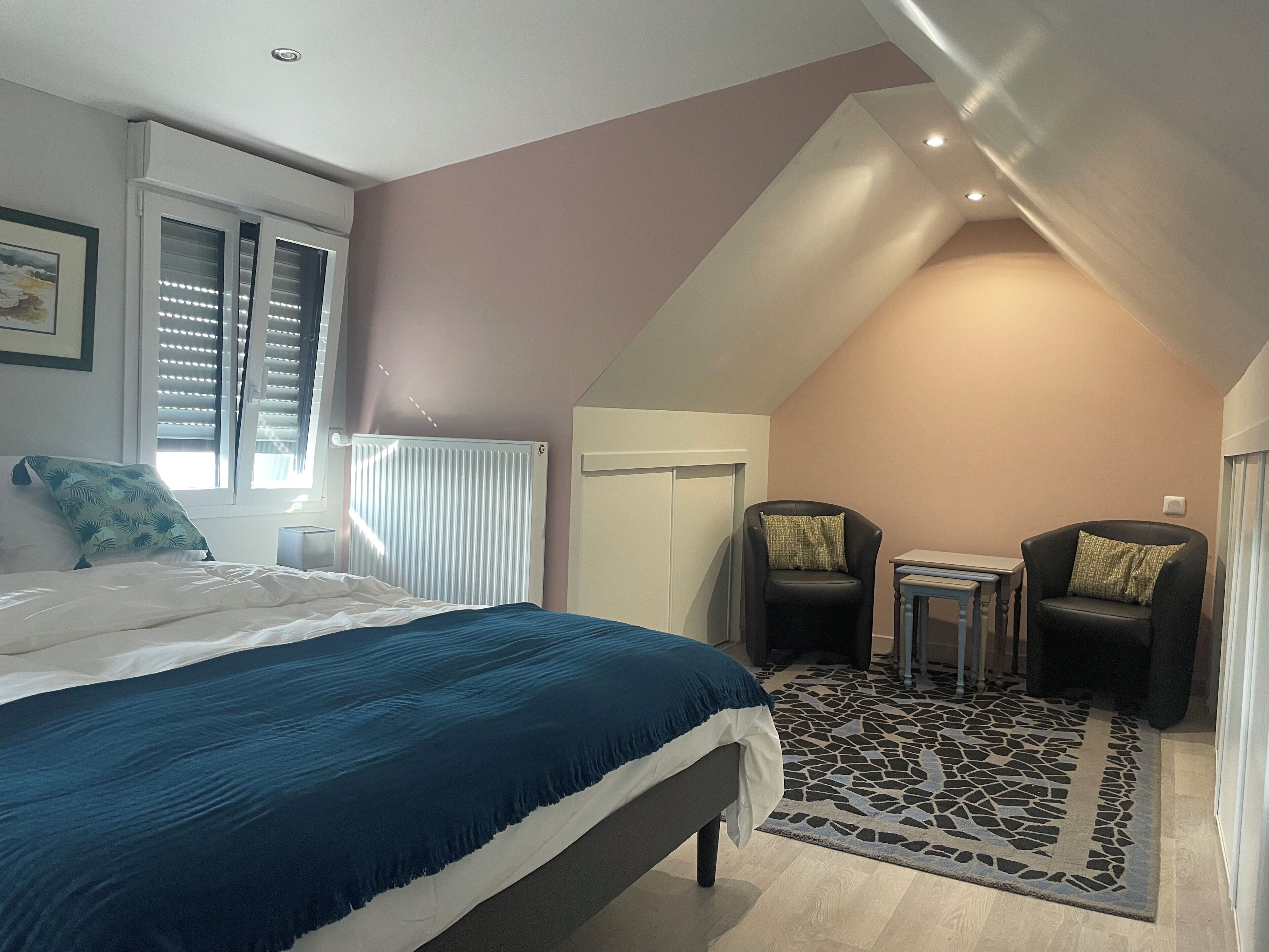 A bedroom with a bed, a radiator, a window with blinds, a small table, two chairs, and a patterned rug.