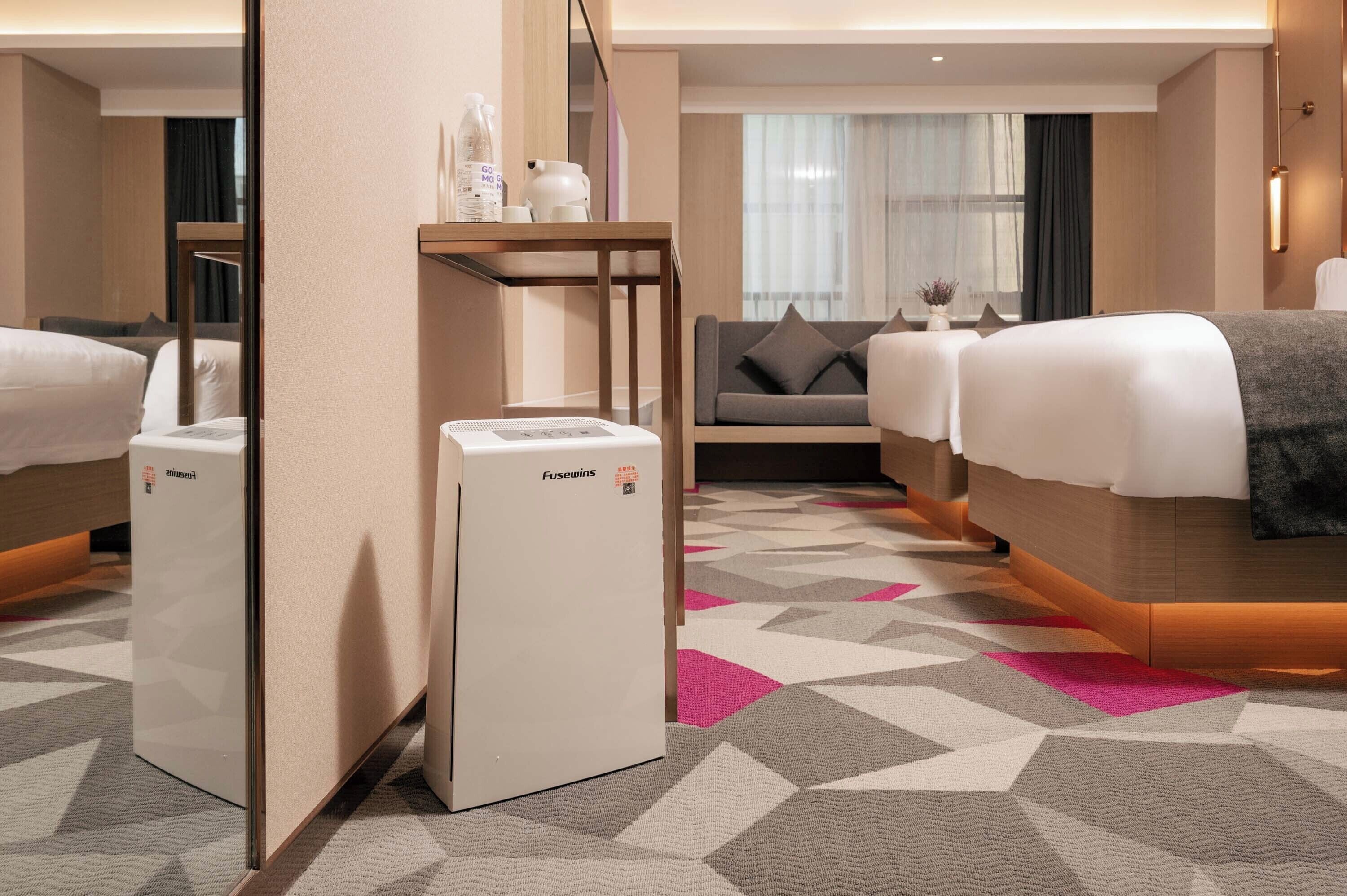 Habitación de hotel con dos camas, un sofá, una mesita con lámpara y un calentador en la pared.