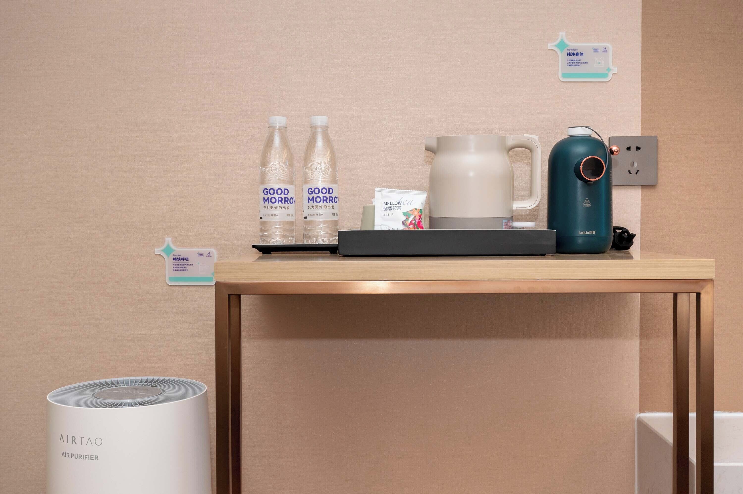 A hotel room with a wooden table holding two bottles of water, a kettle, a coffee maker, and a tea kettle. There is a wall-mounted air purifier and a trash bin.