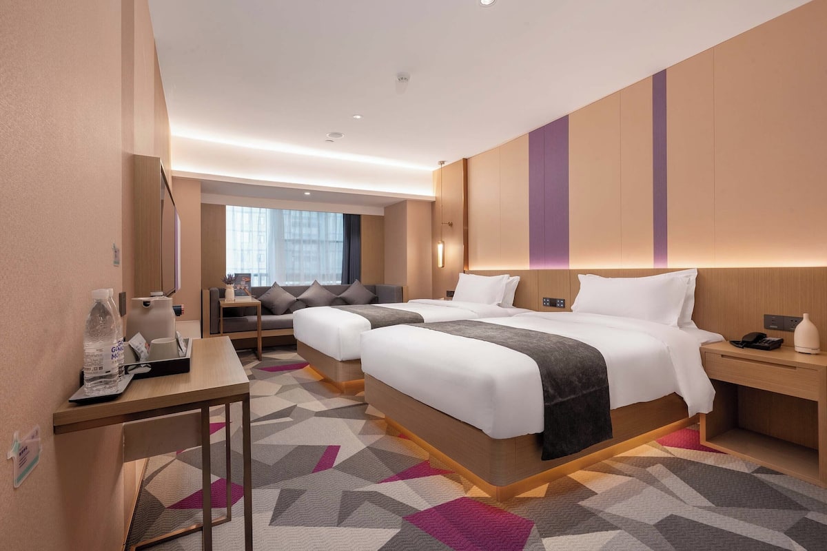 Habitació d'hotel amb un llit gran, una taula de treball i una cadira. Hi ha una finestra amb cortines, una làmpada i una ampolla d'aigua sobre la taula.