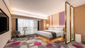 Habitación de hotel con una cama grande, una silla, una mesita y vistas a la ciudad a través de amplias ventanas.