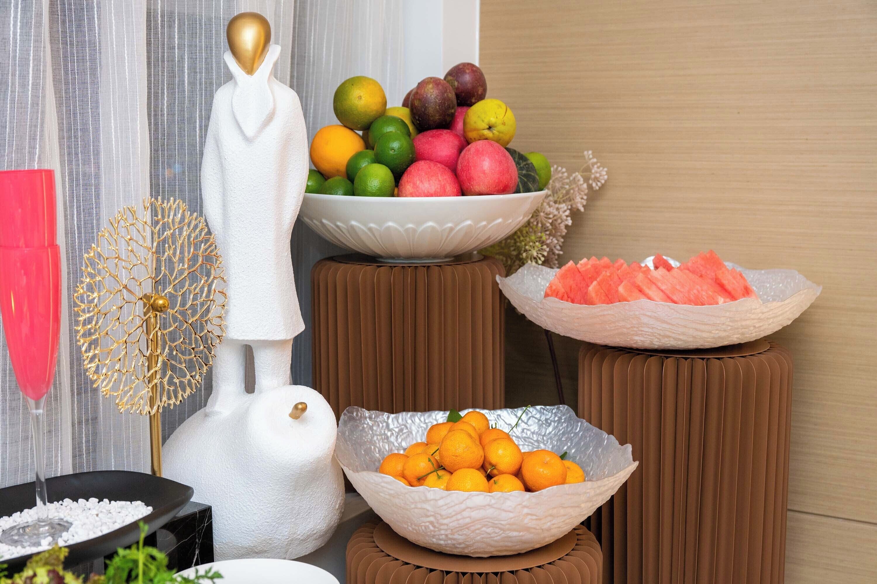 A room with a white statue, a bowl of oranges, a bowl of apples, and a bowl of watermelon.