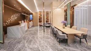 Un moderno vestíbulo de hotel con piso de mármol, una mesa de recepción, una larga mesa de comedor con sillas y un área de descanso con sofás y una mesa de centro.