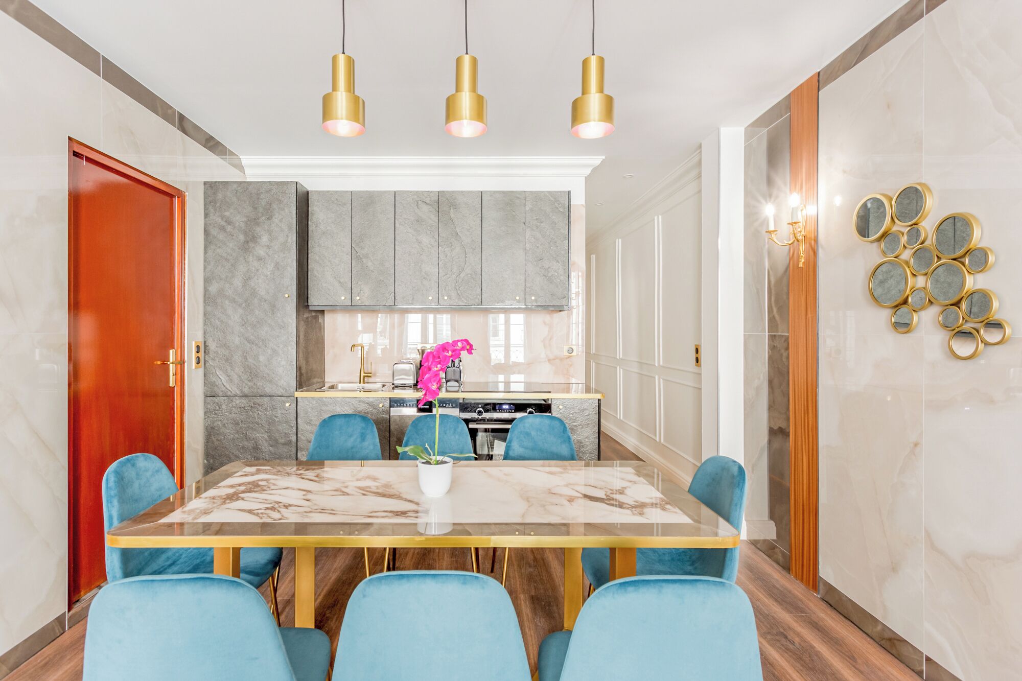 A modern dining area with a marble table, blue upholstered chairs, and a kitchen in the background.