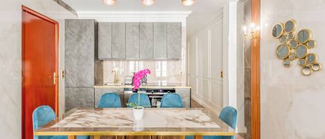 A modern dining area with a marble table, blue upholstered chairs, and a kitchen in the background.