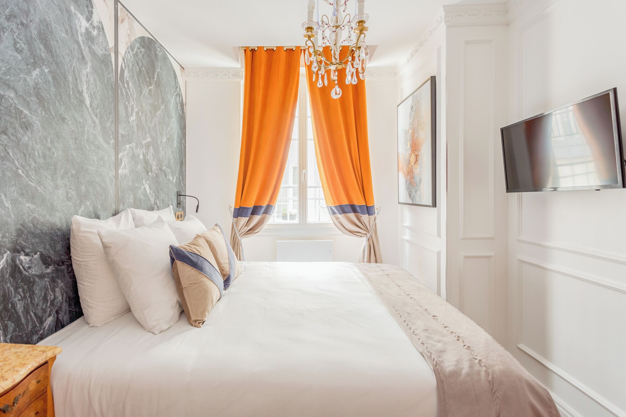 A hotel room with a marble wall, a bed with white bedding, orange curtains, a chandelier, and a flat-screen TV mounted on the wall.