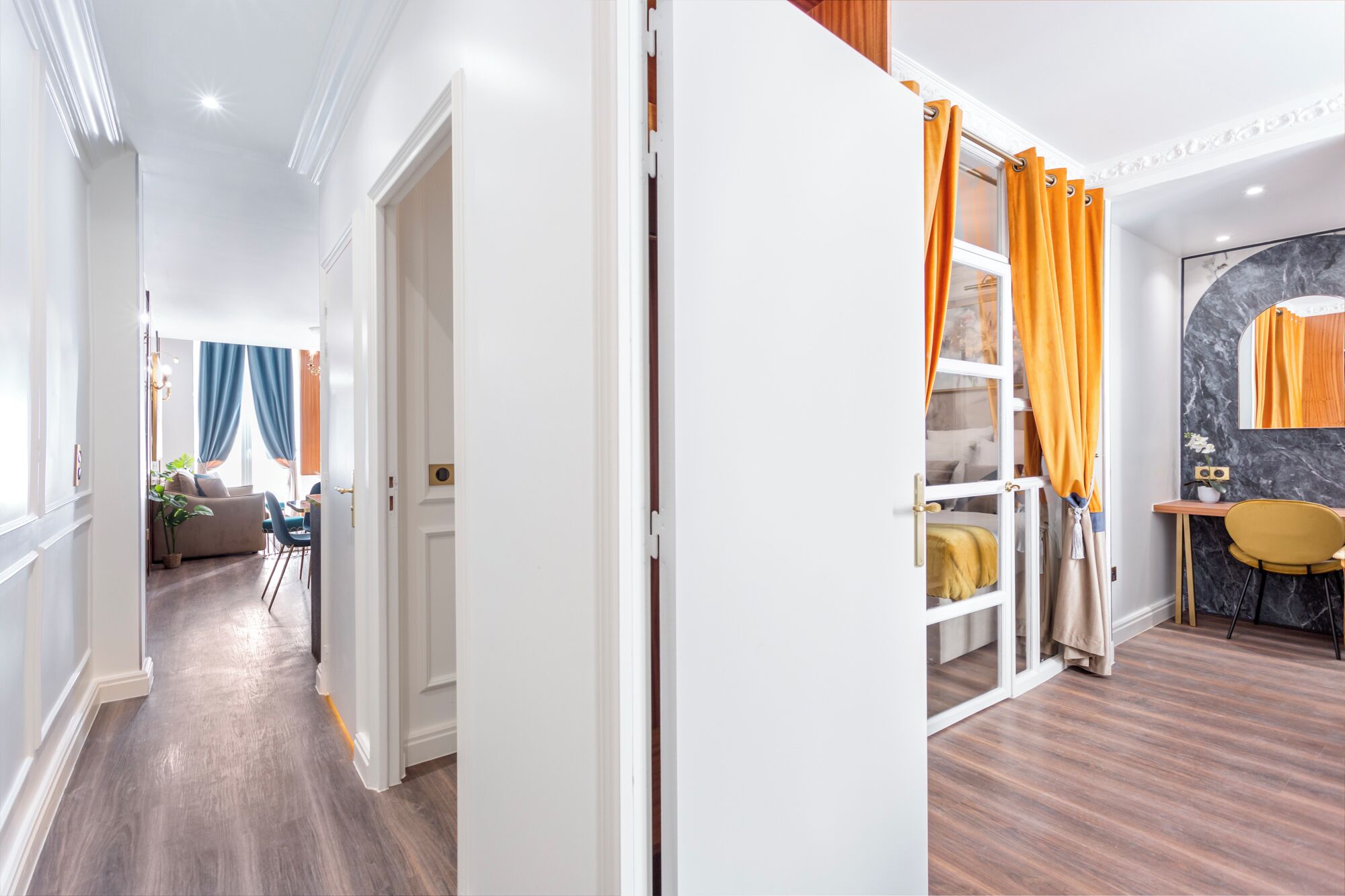 Une chambre d’hôtel avec un plancher en bois, un lit avec une tête de lit jaune, une petite table avec un vase et une fenêtre avec des rideaux orange.