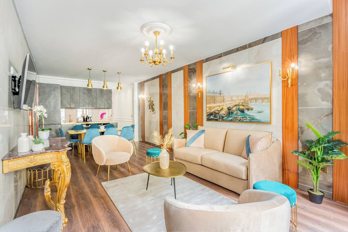 Un salon moderne avec un canapé, des fauteuils, une table basse et un coin repas, la cuisine étant visible au fond.