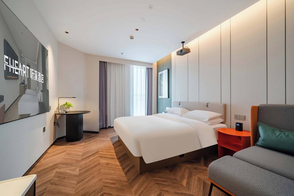 A modern hotel room with a large bed, a seating area with a sofa, a small table with a lamp, and a wall-mounted projector.