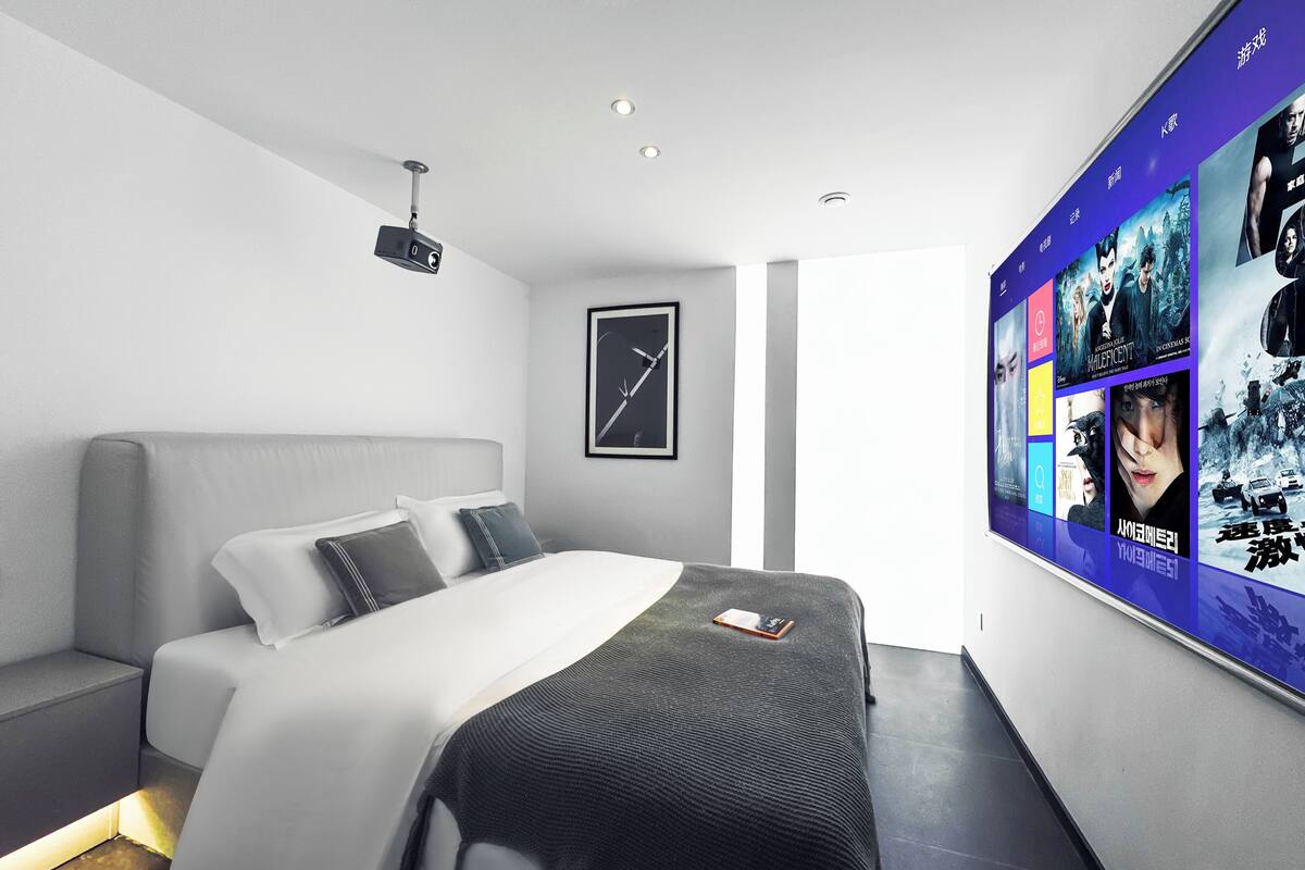 A modern hotel room with a bed, a side table, a projector, and a wall with movie posters.