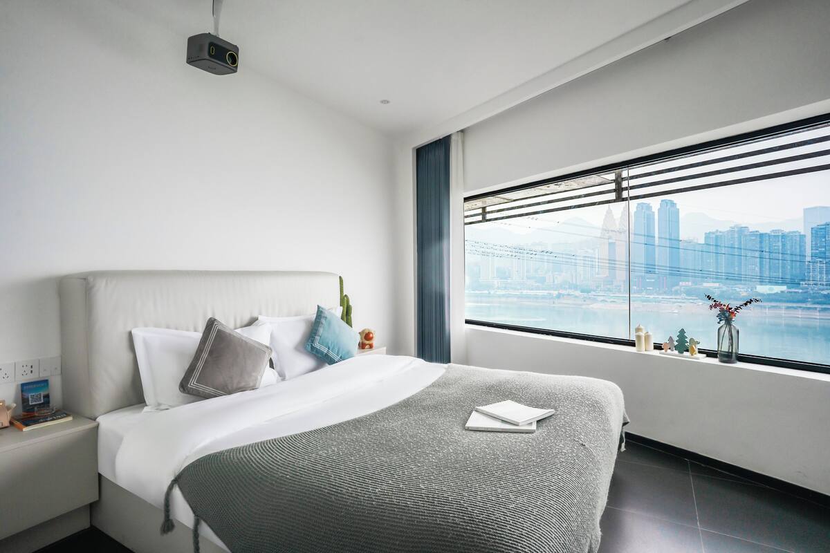 A modern hotel room with a large bed, a view of a city skyline, and a projector mounted on the ceiling.