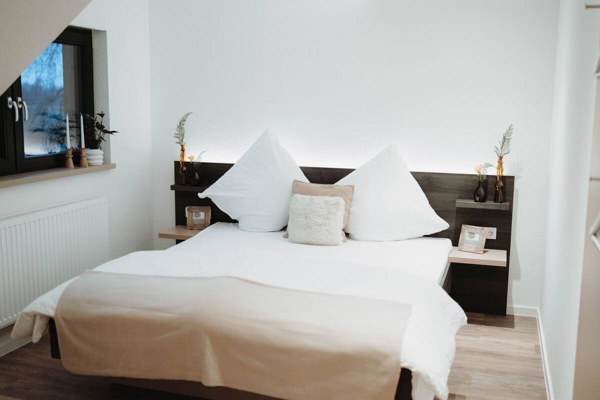 A bedroom with a bed, white bedding, two bedside tables with lamps, and a window with a view.