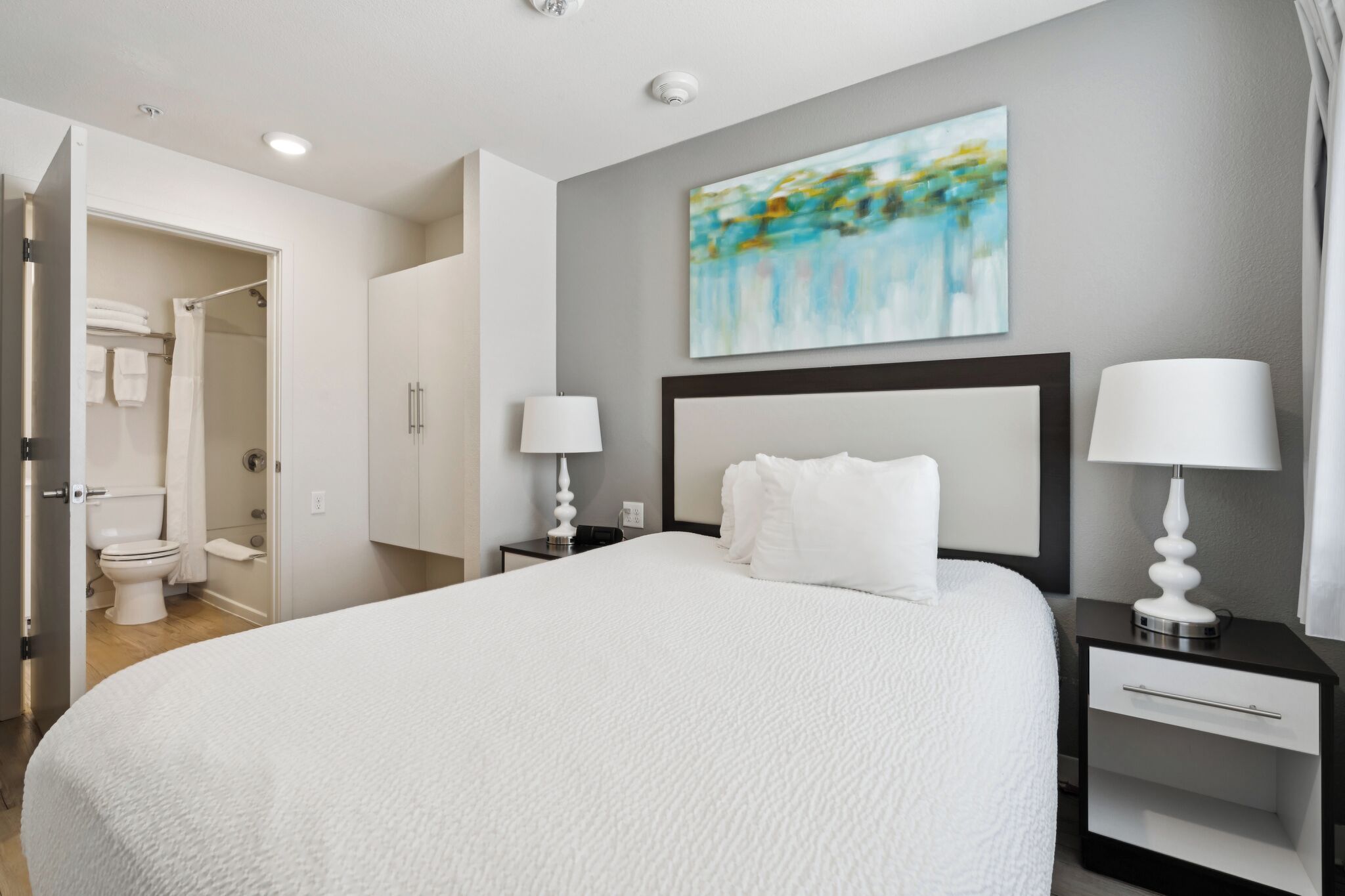 A hotel room with a large bed, two bedside tables with lamps, a headboard, a painting on the wall, and a bathroom visible through an open door.