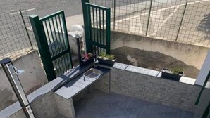 An outdoor patio area with a stone floor, a metal grill, and a green gate. - Reysol Home (Costa Saracena - Castelluccio)