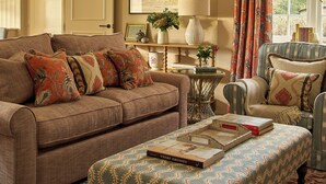 Una sala de estar acogedora con un sofá, un sillón y un reposapiés, una mesa con una lámpara y una ventana con cortinas.