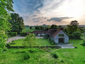 Une maison traditionnelle en pierre, avec un toit de chaume, entourée de jardins bien entretenus et d’un paysage rural.