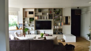 Een moderne woonkamer met een grote flatscreen televisie, inbouwplanken met boeken en decoratieve objecten, een comfortabele bank en uitzicht op het buitenlands landschap door een raam.