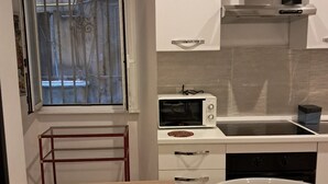 Eine kleine Küche mit weißen Schränken, einer Mikrowelle und einem Spülbecken. Es gibt ein Fenster mit Metallgitter und einen Holztisch mit einer Schüssel.