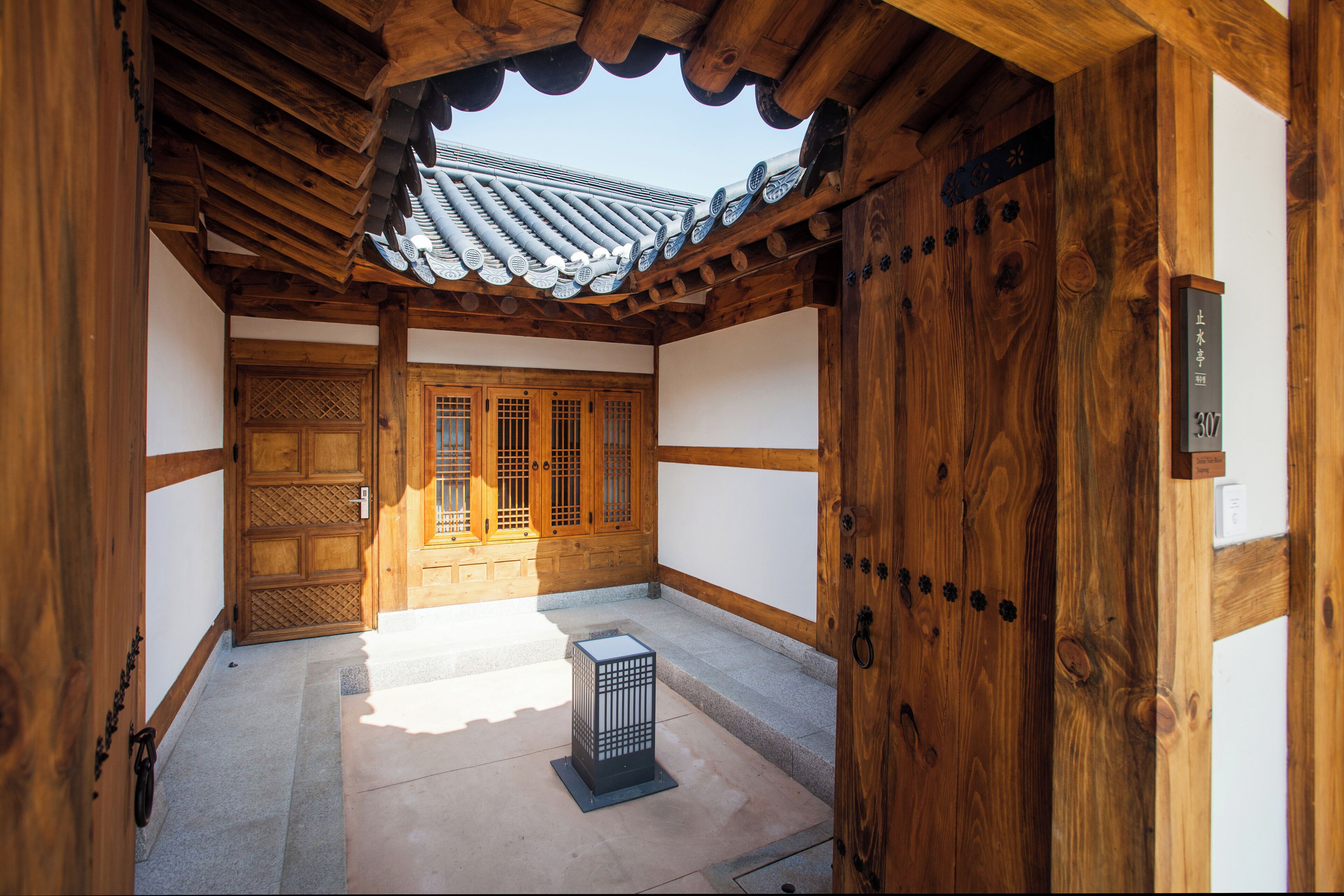 A traditional wooden building with a tiled roof, a wooden door with a number plate, and a small black object on the ground.