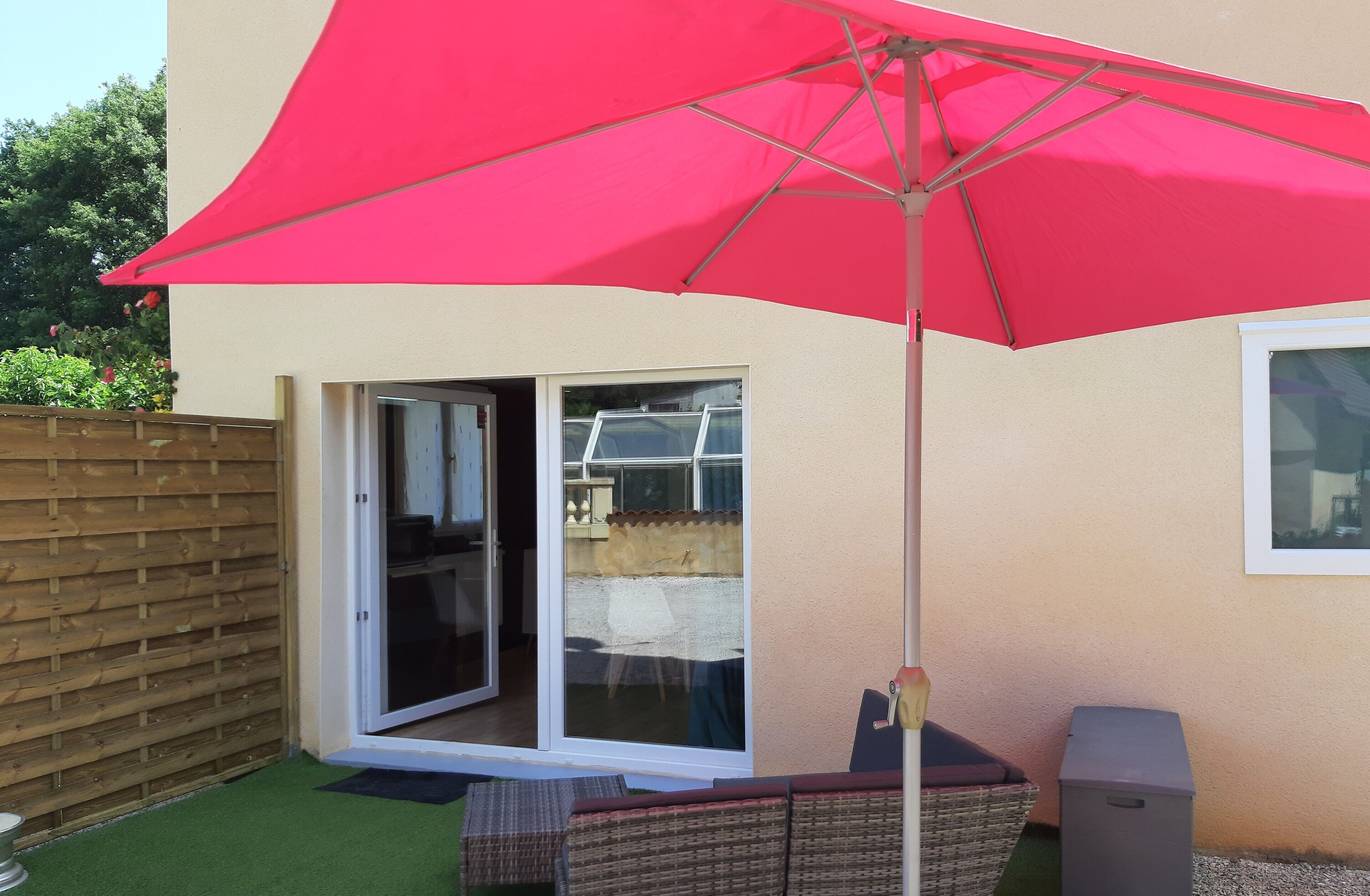Eine Terrasse mit rotem Sonnenschirm, einer Glasschiebetür, einer Korbsessel-Garnitur und einem Mülleimer.