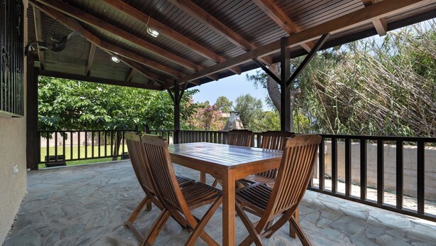 Une terrasse couverte avec une table et des chaises en bois, un sol en pierre et une vue sur la verdure.