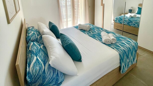 A double bed with a blue and white patterned bedspread, two white pillows, and a wooden headboard. There is a large mirror on the wall, a window with sheer curtains, and a hanging pendant light. - NEW! 2 Bedrooms, 2 Bathrooms, Free Car Box 100m from the sea (Alba Adriatica)