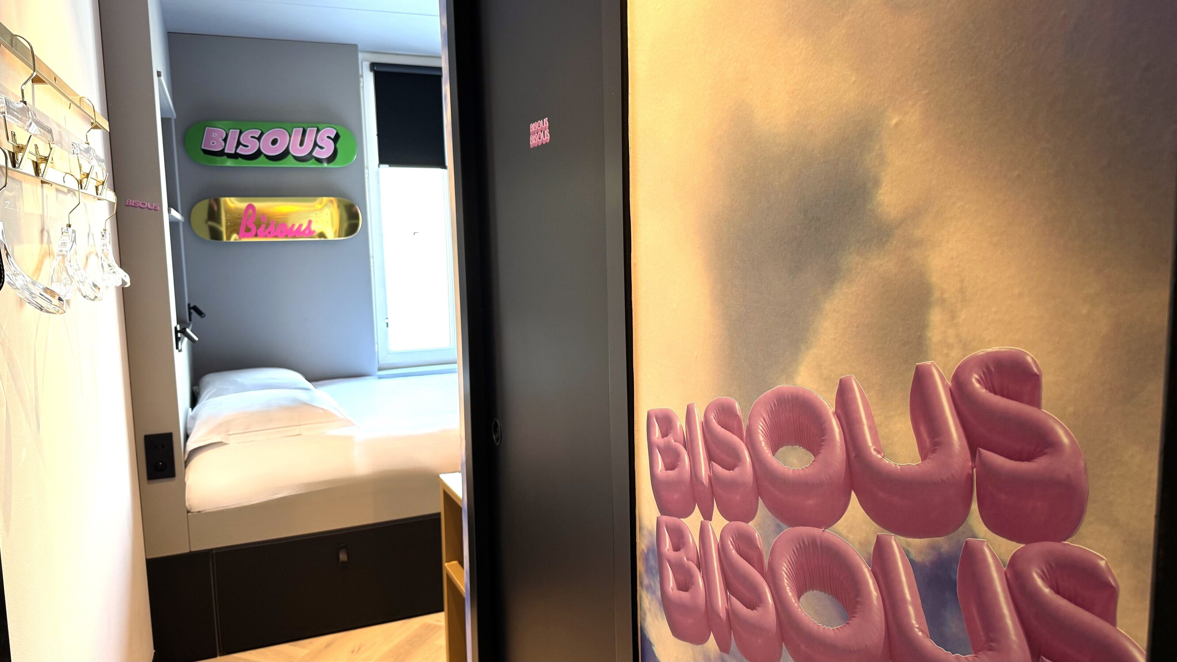 A hotel room with a bed, a window, and a wall with a 'BISOU' sign.