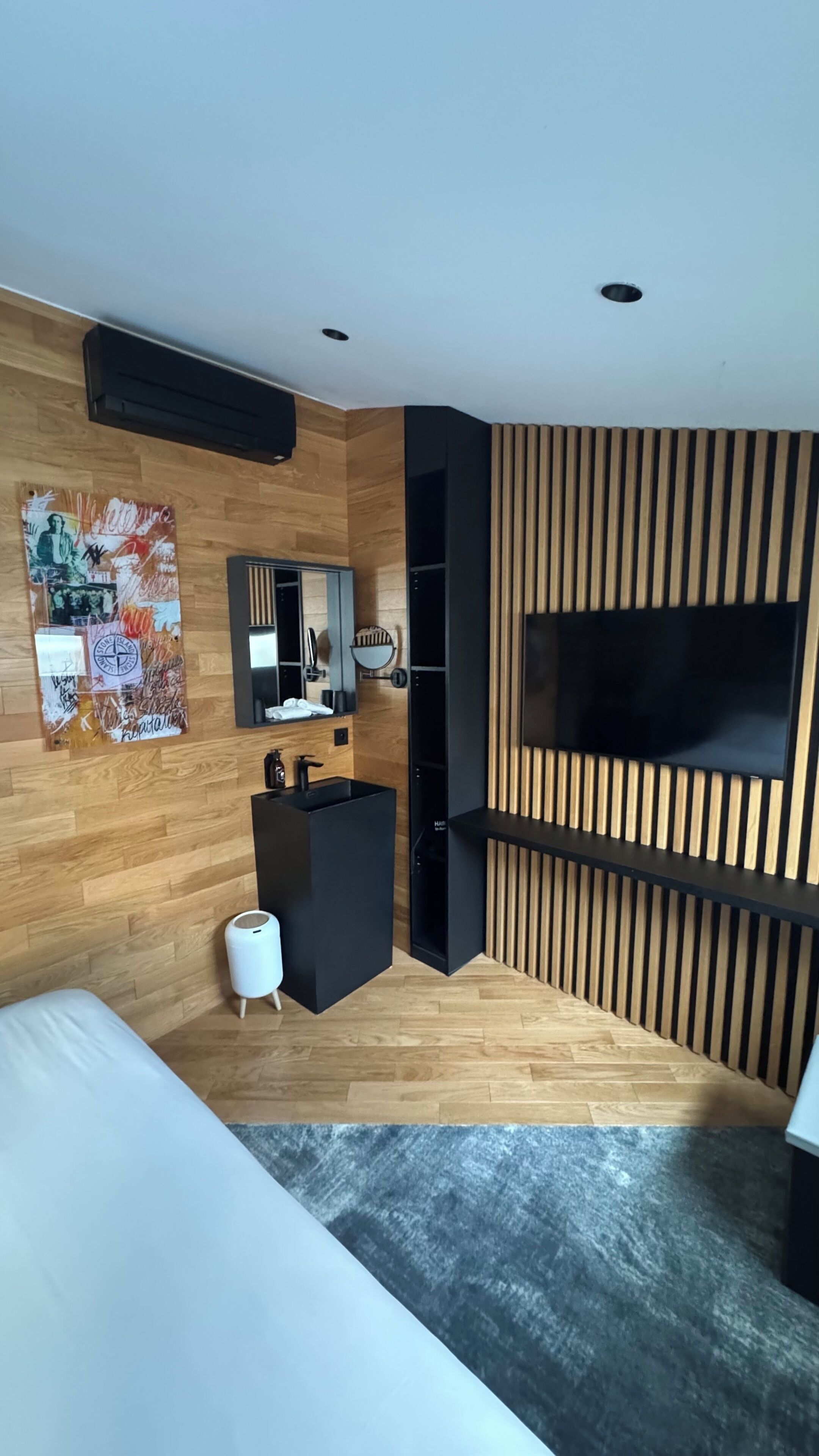Ein modernes Hotelzimmer mit Holzboden, einem großen Bett, einem schwarzen Schminktisch mit Spiegel, einem weißen Nachttisch, einem schwarzen Fernseher, der an einer Wand aus Holzpaneelen befestigt ist, und einem großen Fenster.