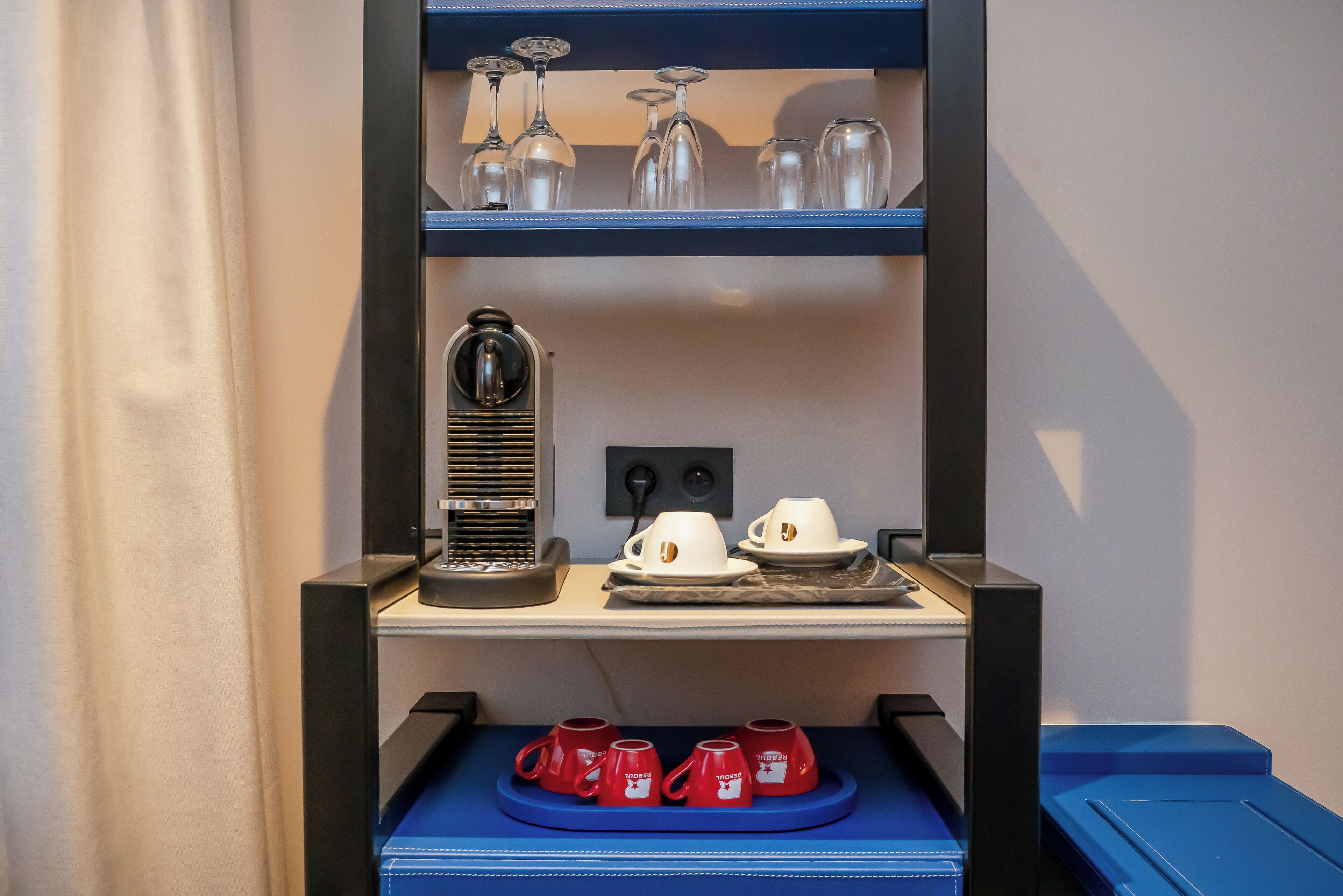 Een hotelkamer met een blauwe plank met wijnglazen, een koffiezetapparaat en rode mokken.
