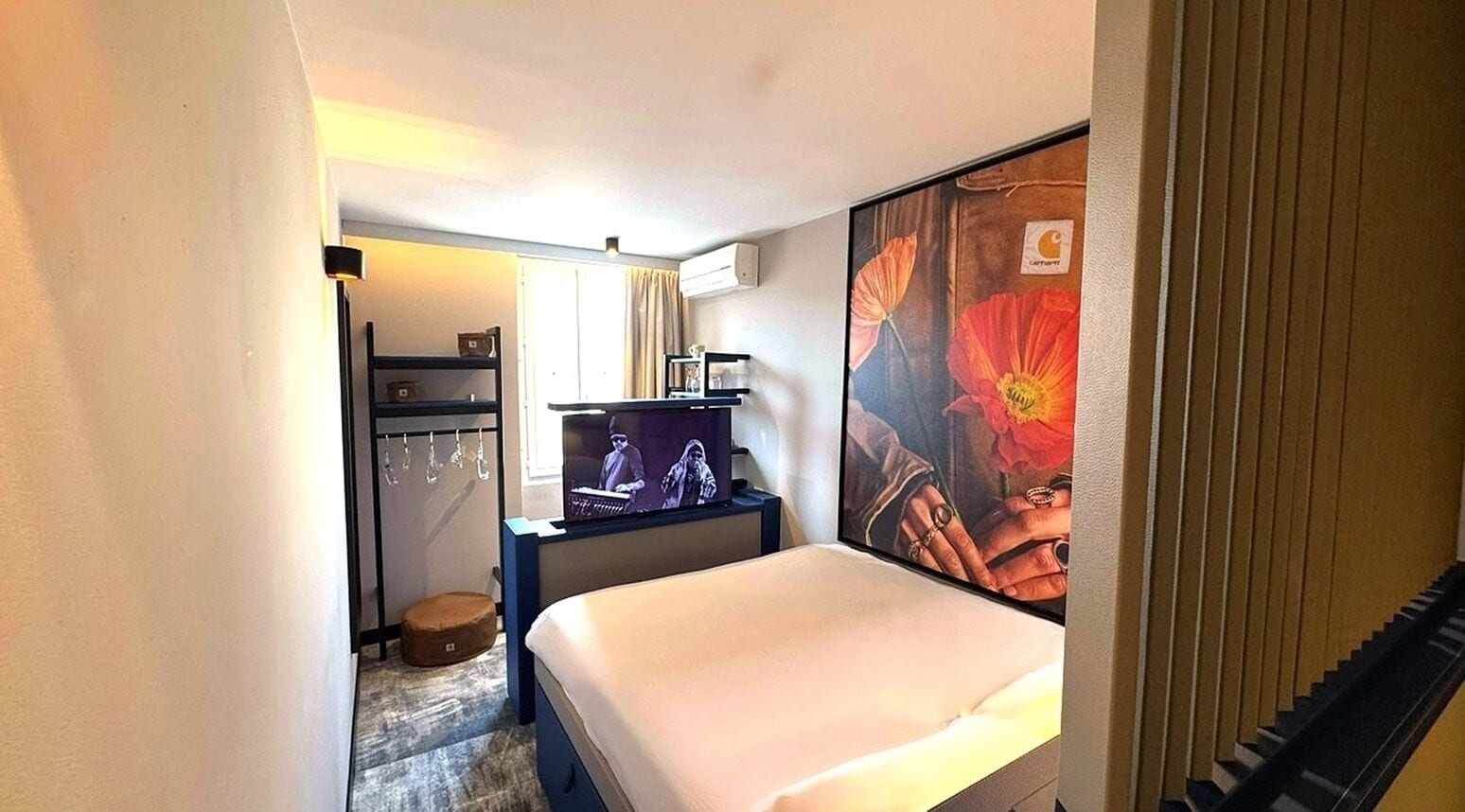 A hotel room with a bed, a television, a desk with a chair, and a large floral artwork on the wall.