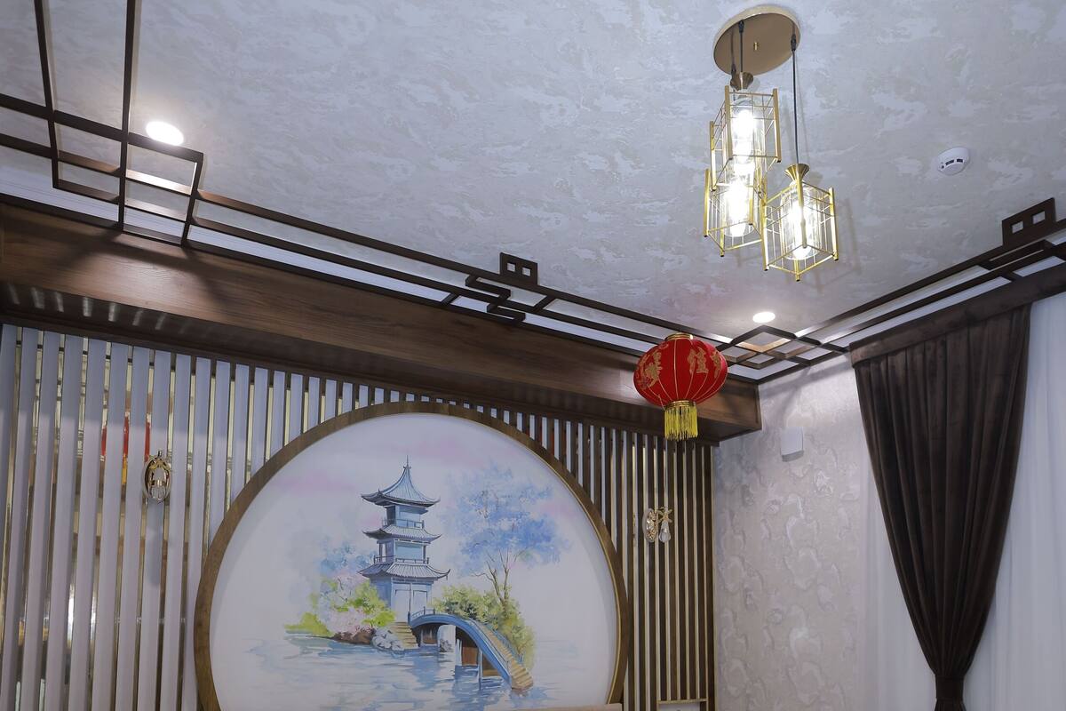 Une chambre à coucher comprenant un lit, des tables de chevet, une commode, une lampe fixée au mur, un luminaire au plafond, un élément décoratif mural et une fenêtre avec des rideaux.
