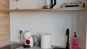 Et køkken med vask, elkedel, kaffemaskine, en rulle køkkenrulle, opvaskesæbe og et glas på en hylde.