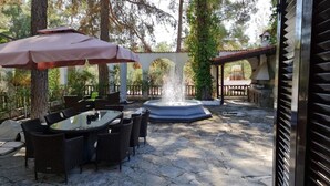 An outdoor patio area with stone flooring, a round table with chairs, a fountain, and umbrellas. - Charming Villa in Moniatis, Cyprus - Private Pool & Garden (Saittas)