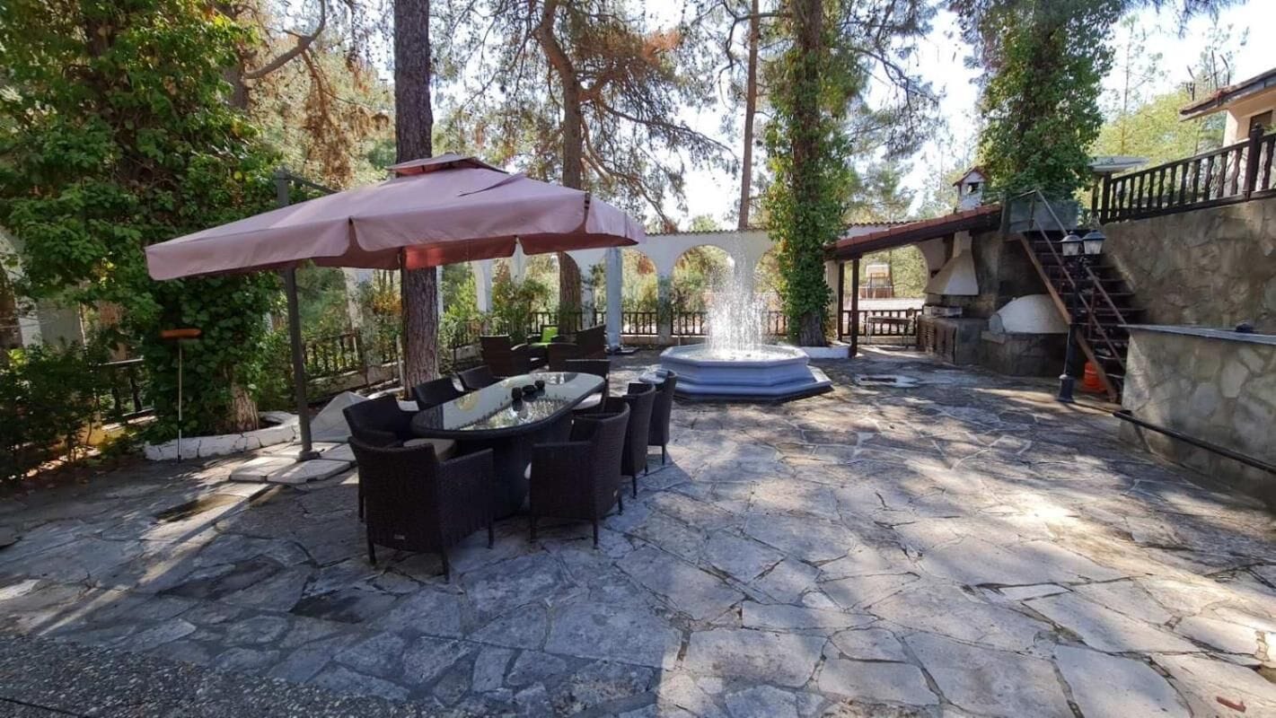 An outdoor patio with stone flooring, a round table with chairs, a large umbrella, and a fountain.