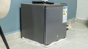 A compact, gray portable heater with a remote control on top, placed on a tiled floor against a blue wall. - The Den Soroti Red (Soroti)