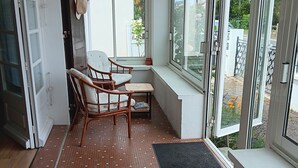 Un petit balcon fermé, agrémenté de chaises en osier, d’une petite table et offrant une vue sur un jardin grâce à une porte vitrée.