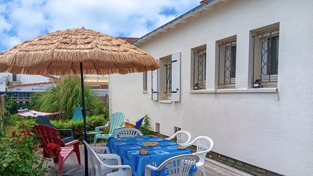 Une terrasse extérieure avec une table et des chaises, un parasol en chaume et un bâtiment blanc doté de fenêtres.