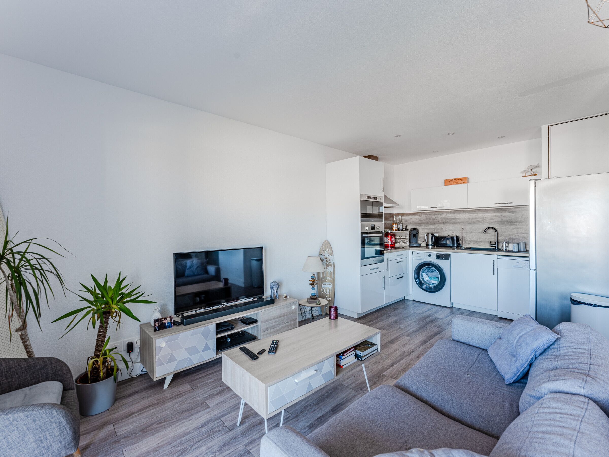 A modern living room with a flat-screen TV, a coffee table, and a sofa. Adjacent is a compact kitchen with a washing machine, oven, and microwave.