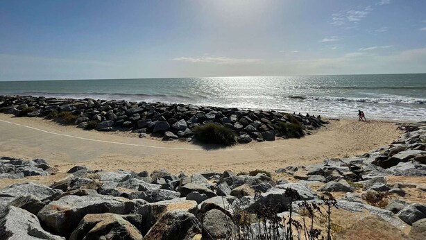 Ein Strand mit einem steinernen Wellenbrecher, einem Sandweg und dem Meer im Hintergrund.