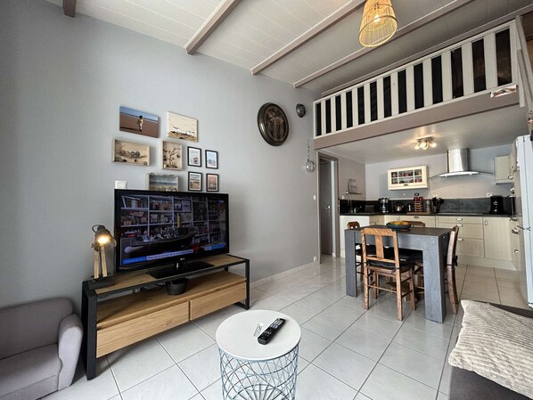 A modern living room with a flat-screen TV, a dining area with a table and chairs, and a kitchen with a stove and cabinets.